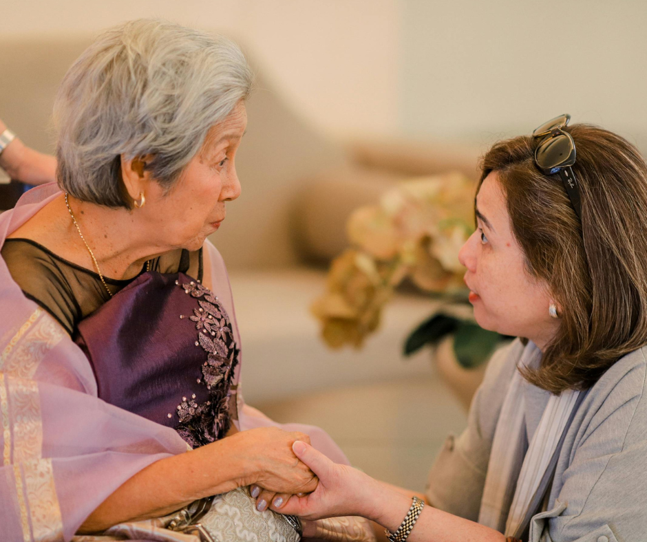 A younger woman is talking to an elderly woman whilst holding her hand.